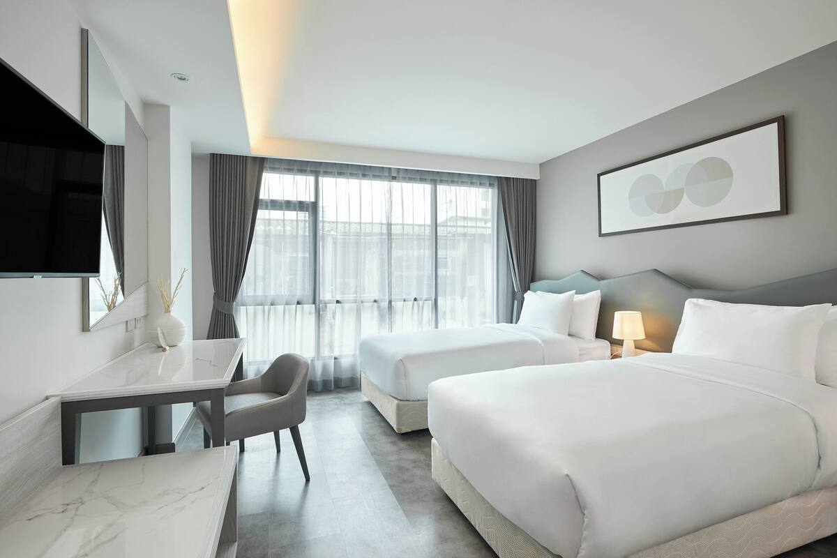 Una habitación de hotel moderna con una cama grande, una cama más pequeña, un escritorio con silla, un televisor de pantalla plana y un ventanal con cortinas.