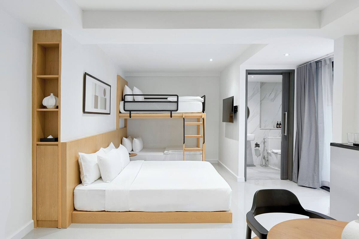 Una habitación de hotel moderna con una cama litera, una mesita de noche de madera, un escritorio y un baño visible a través de una puerta abierta.