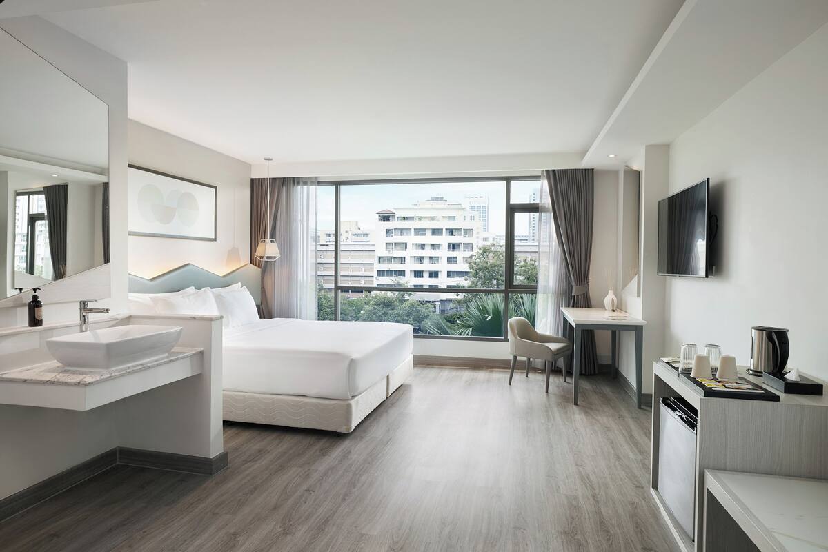 Una habitación de hotel moderna con una cama grande, un escritorio, una silla, un televisor y vistas de la ciudad a través de amplios ventanales.