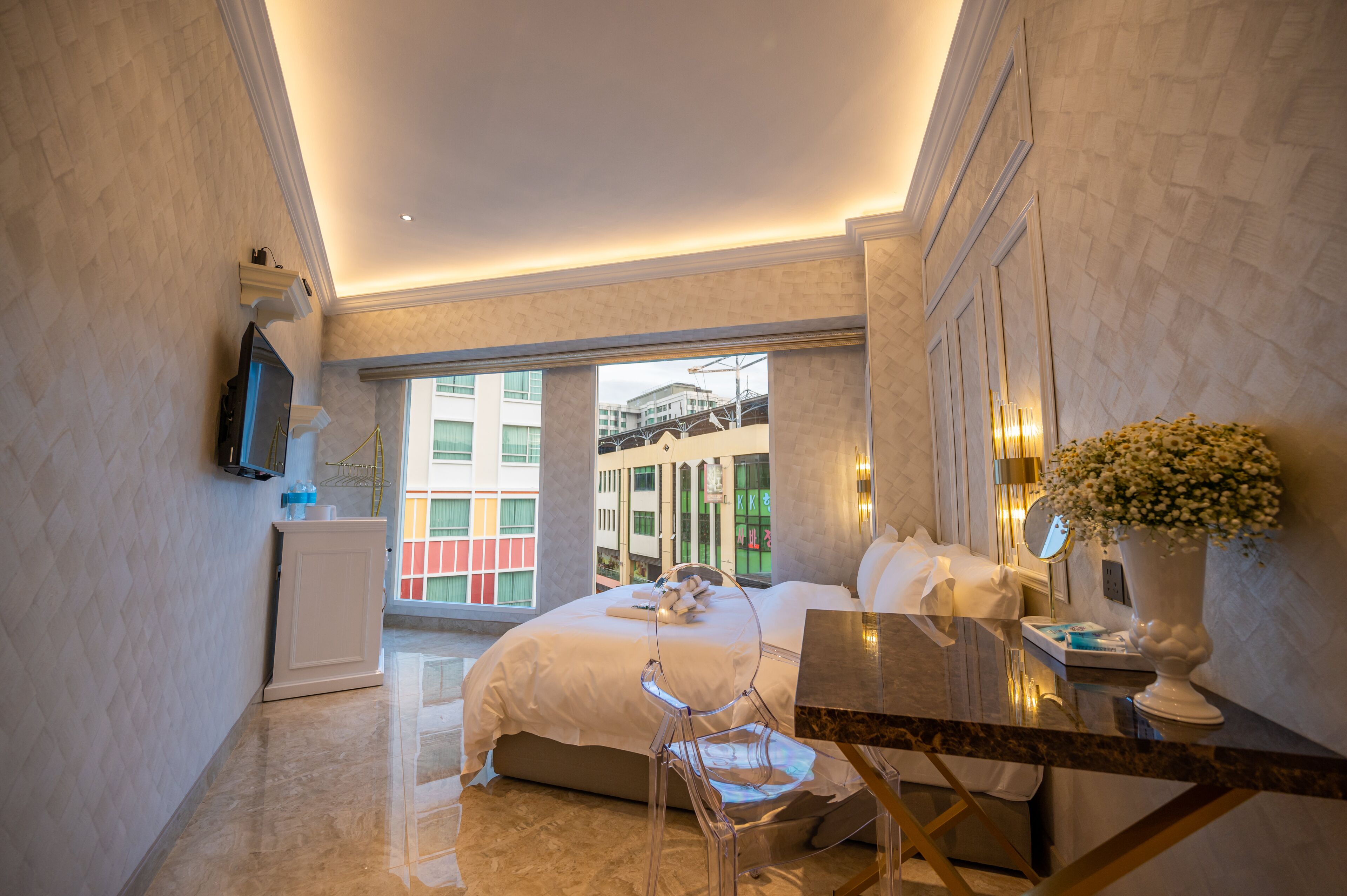 Quarto de hotel com cama, escrivaninha, cadeira, televisão, um vaso com flores e janela com vista para edifícios.
