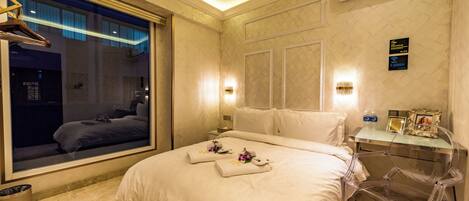 A hotel room with a large bed, a bedside table, a clear chair, a window with a view of the outside, and an air conditioning unit.