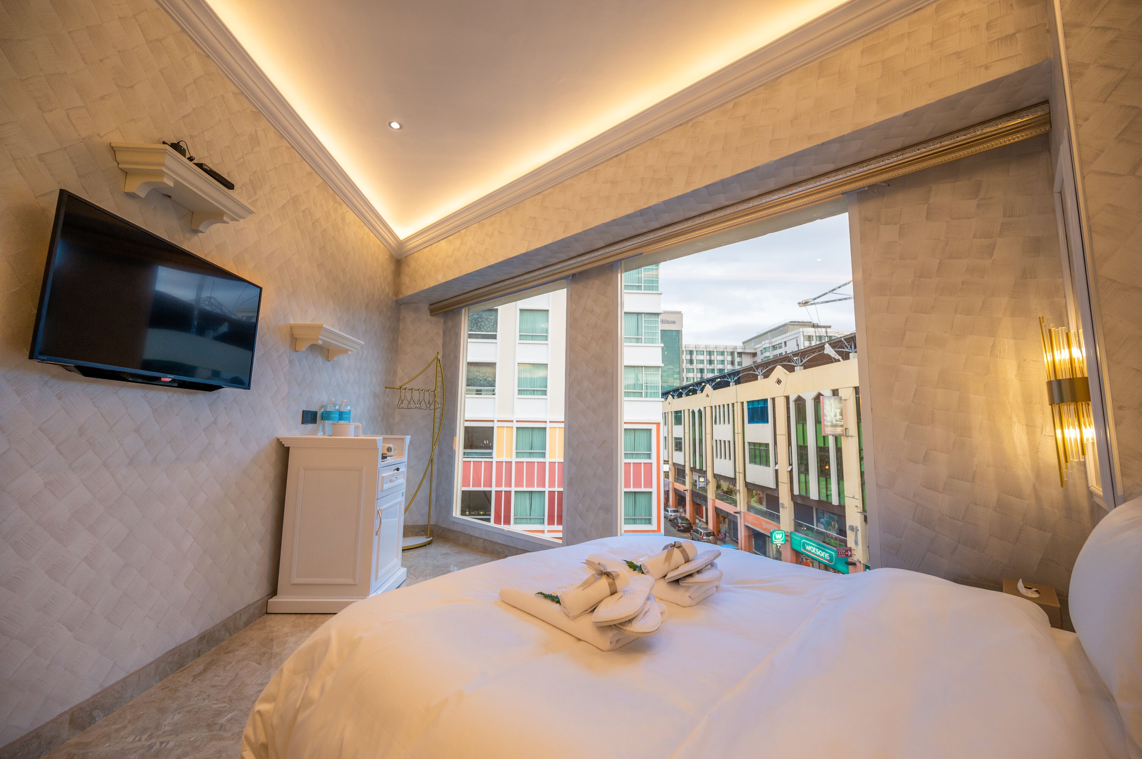 A hotel room with a bed, a TV mounted on the wall, a window with a view of buildings, and a cabinet.