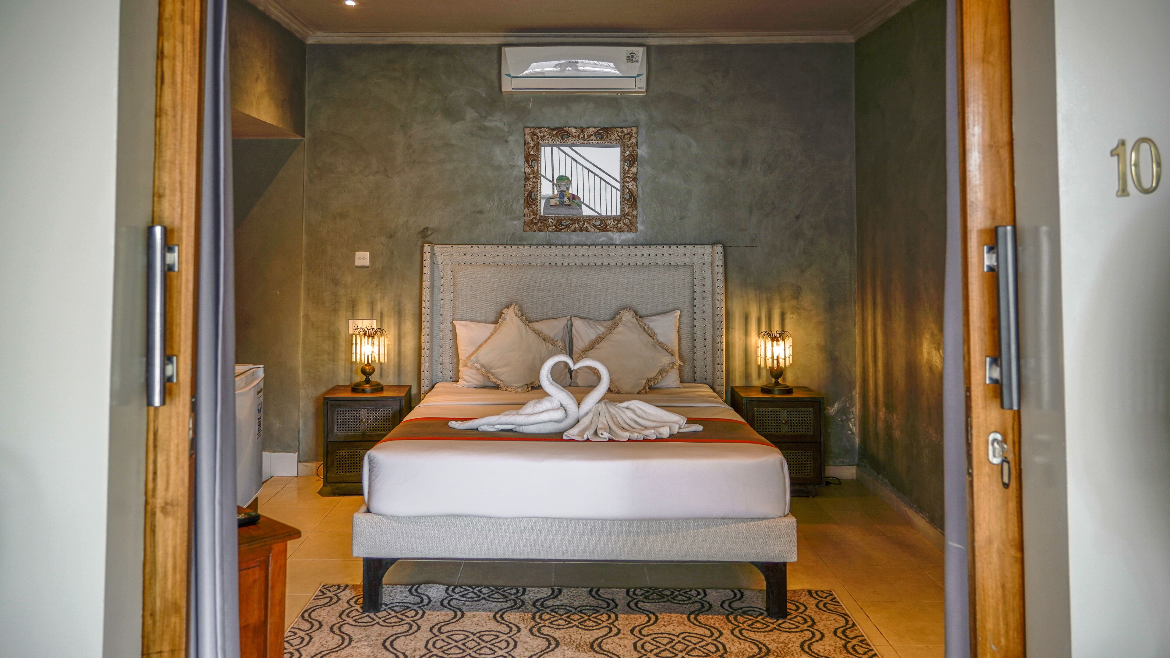 A neatly made bed with a heart-shaped towel arrangement, a large mirror above the headboard, and two bedside lamps.