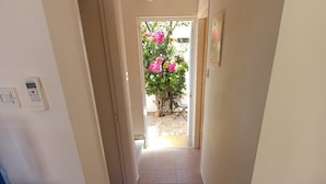 Een smalle gang met betegelde vloer, een plafondlamp en een deur die uitkomt op een tuin met roze bloemen.