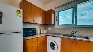 Een kleine keuken met houten kastjes, een magnetron, een wasmachine en een raam boven de gootsteen.