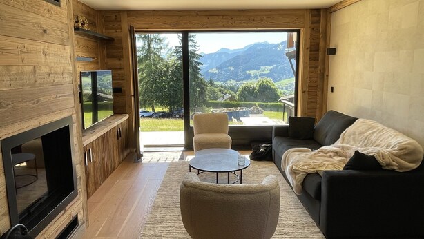 A modern living room with a large window, a sofa, a coffee table, and a TV mounted on the wall.