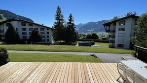 A wooden deck with a view of two modern buildings, a grassy area, and distant mountains.