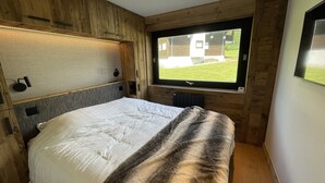 A bedroom with a bed, wooden headboard, a window with a view of a building and lawn, and a wall-mounted light fixture.