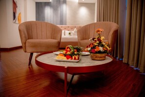 A room with two beige armchairs, a round table with a fruit platter and a floral arrangement, and a bed with a white headboard in the background. - Lavendish Hills Kandy (Kandy)
