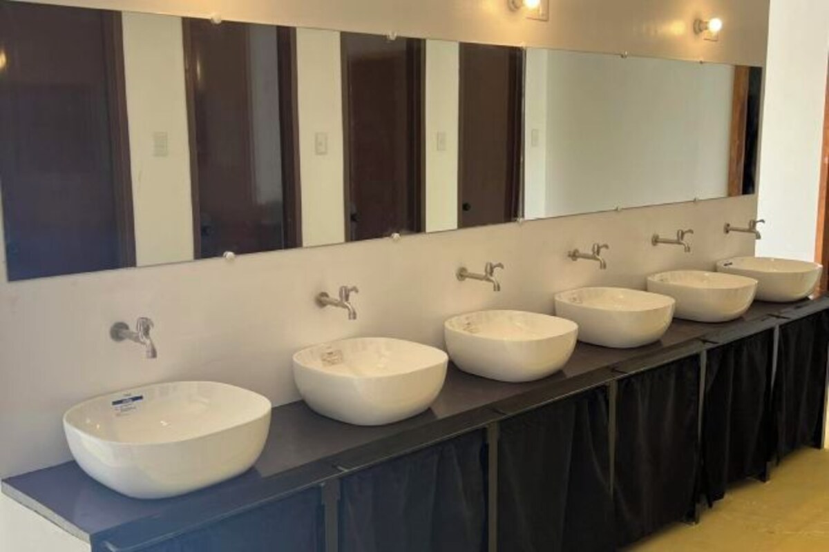 A row of white vessel sinks with individual faucets, a large mirror above, and a black curtain below.