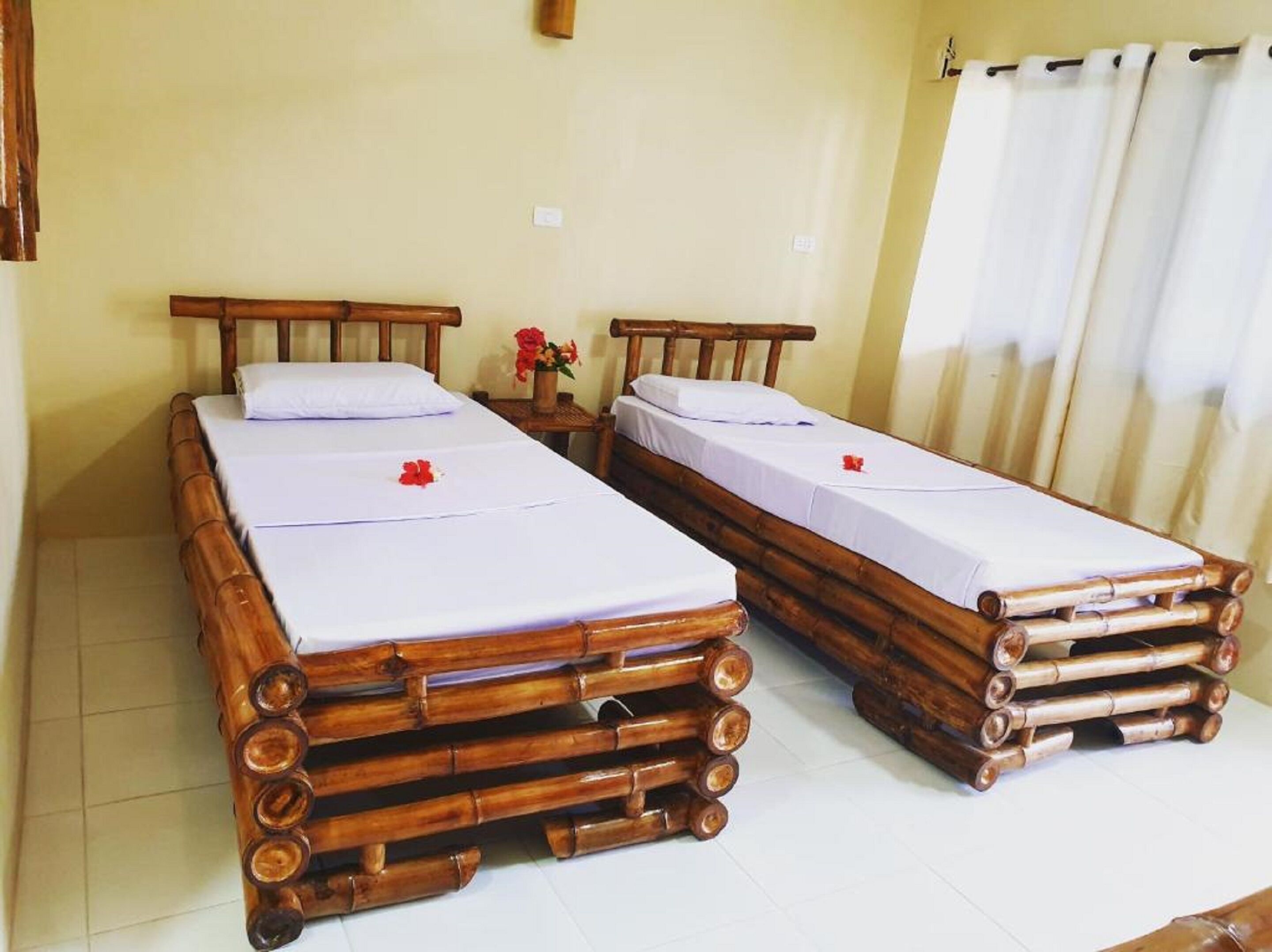 Two wooden beds with white linens and red flowers, a small table with a vase of flowers, and a window with curtains.