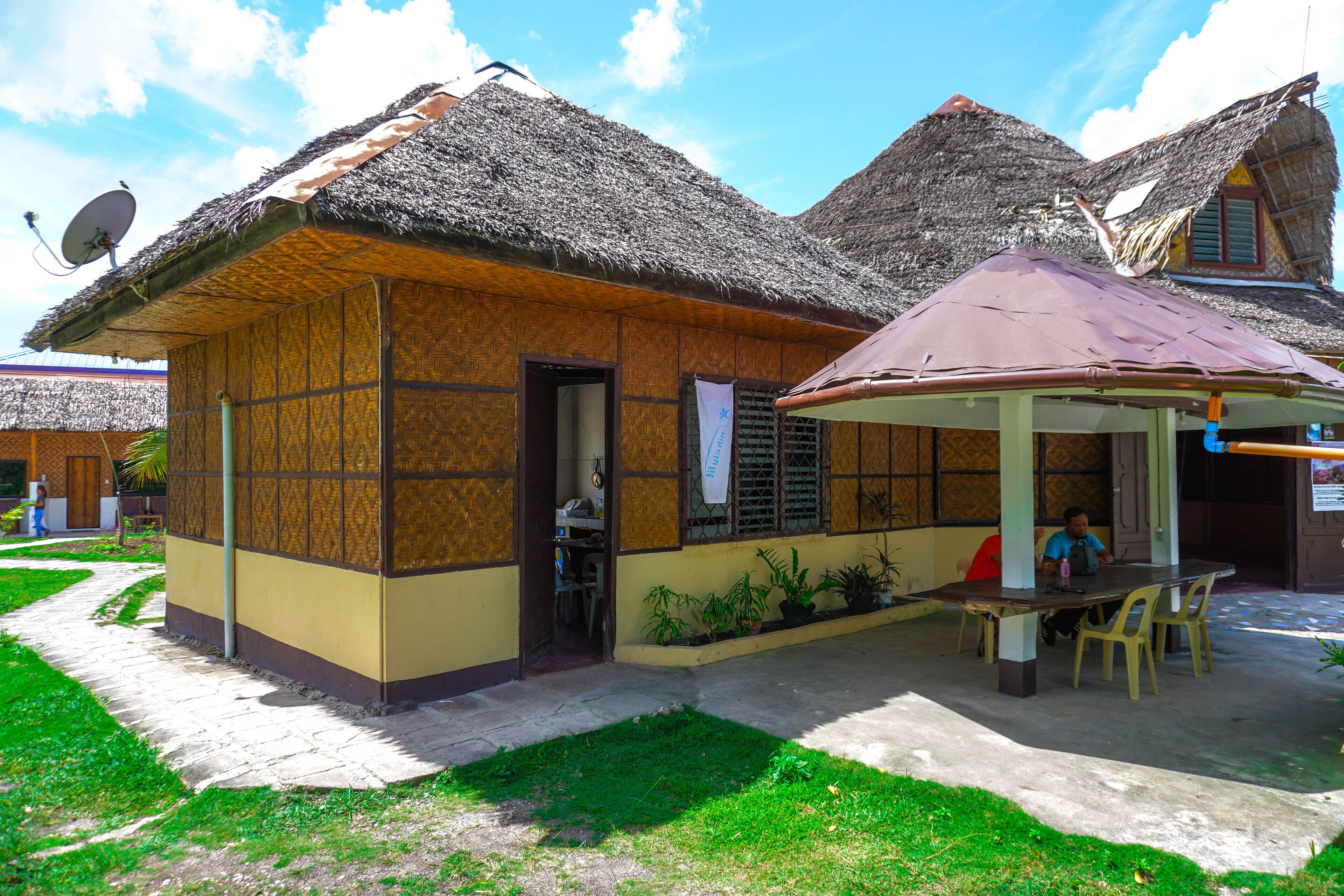 Una casa tradicional con techo de paja y una pequeña veranda, una antena parabólica y una mesa con sillas.