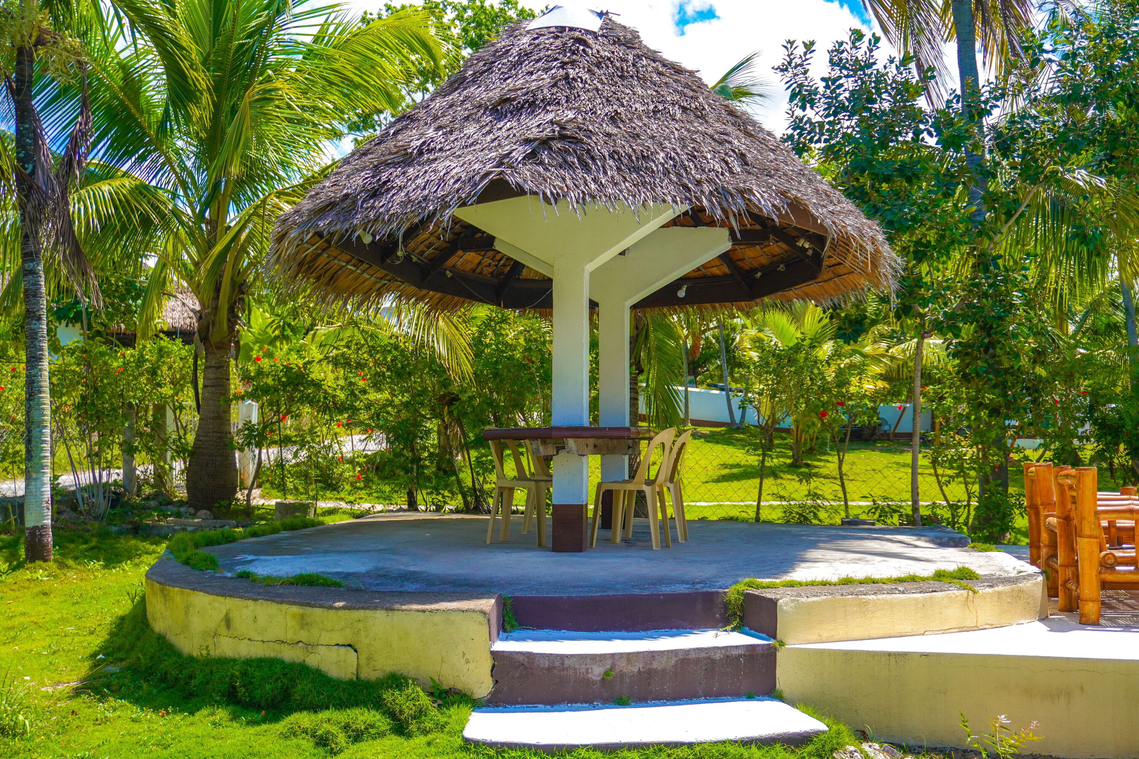 A thatched-roof gazebo with a table and chairs, surrounded by lush greenery and palm trees.