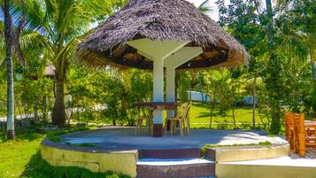 A thatched-roof gazebo with a table and chairs, surrounded by lush greenery and palm trees.