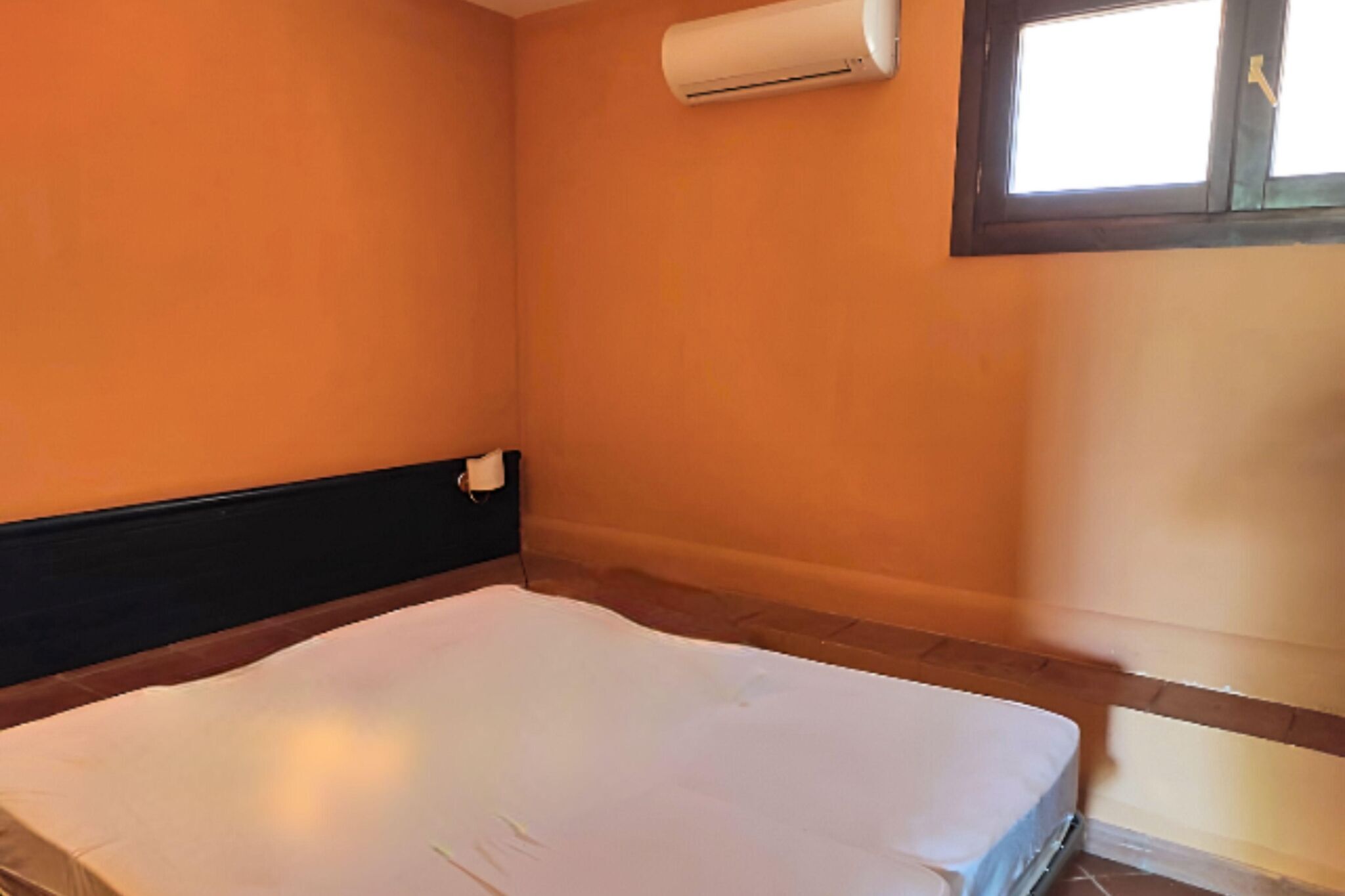 A room with an air conditioning unit mounted on the wall, a window with a view, and a bed with a white mattress.