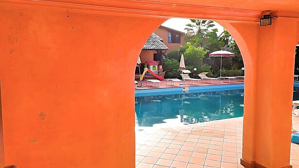 Zona de piscina exterior con solárium de azulejos y área de descanso cubierta.