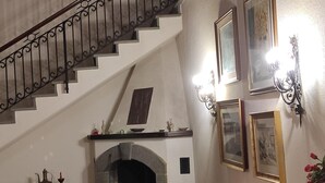 Una escalera con barandilla de hierro forjado, una chimenea con arco de piedra y marcos de cuadros en la pared.