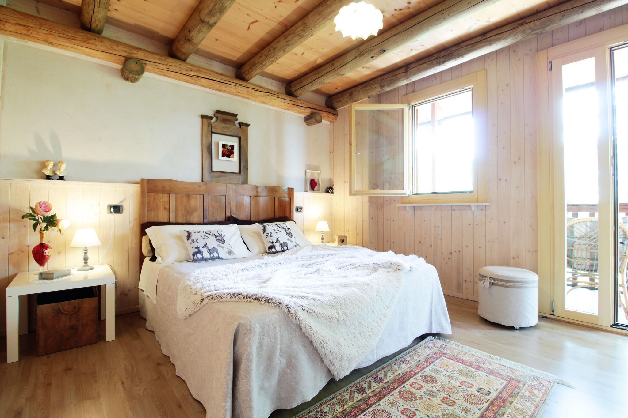 A cozy bedroom with a wooden ceiling, a large bed with a white bedspread, two bedside tables with lamps, a small round ottoman, and a large window with a view of the outside.