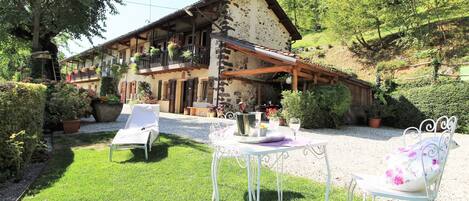 A rustic chalet with a balcony, surrounded by lush greenery and a gravel path.