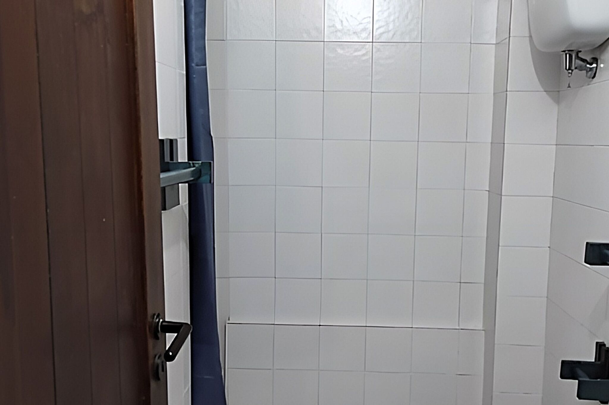 Une salle de bain avec des murs recouverts de carreaux blancs, un rideau de douche bleu et un lavabo blanc avec un robinet chromé.