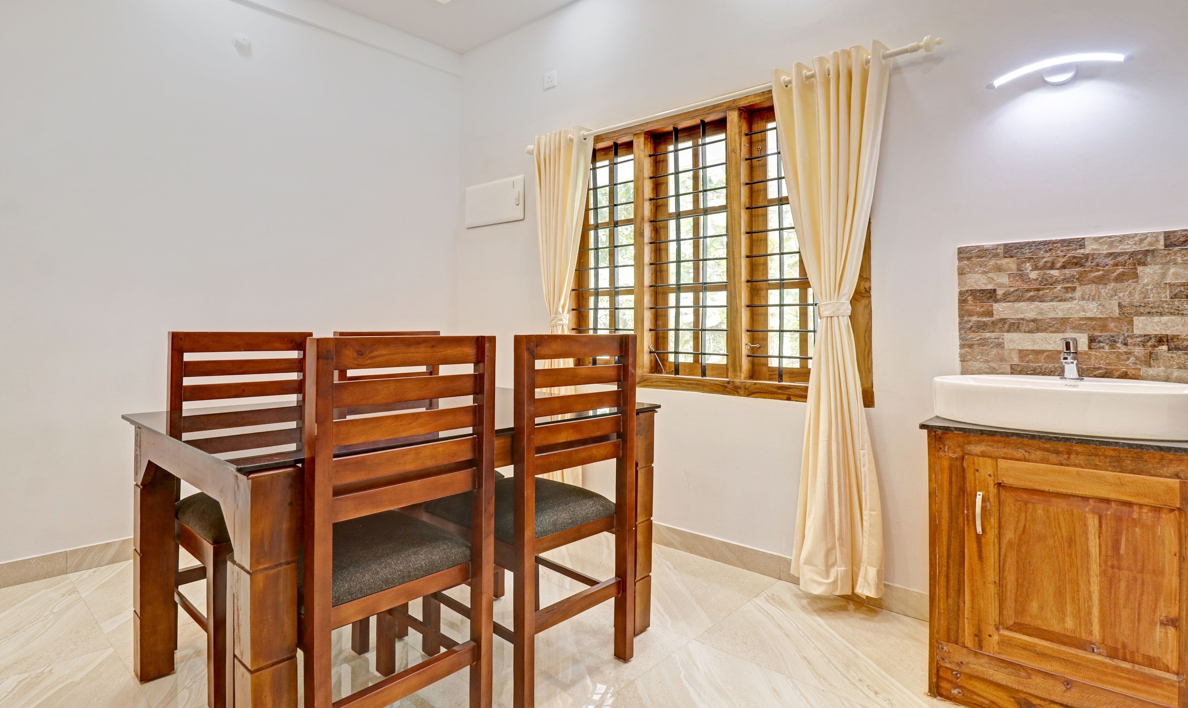 A dining area with a wooden table and chairs, a window with curtains, and a kitchen with a sink and tiled backsplash.