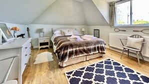 Une chambre avec un plafond en pente, un lit recouvert d’une couverture à rayures, une table de chevet avec une lampe, une chaise et une fenêtre avec des rideaux.