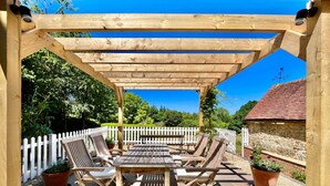 Une pergola en bois avec une table et des chaises, entourée d’une clôture blanche en bois, se trouve dans un jardin agrémenté d’un mur de pierre et, au fond, d’un bâtiment au toit rouge.