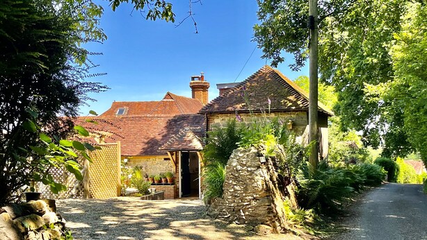 Une maison de campagne en pierre, avec un toit en tuiles, entourée d’une végétation luxuriante et bordée d’une allée en gravier.