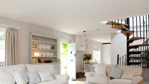 A spacious living room with two white sofas, a wooden coffee table, and a bookshelf filled with decorative items. A staircase with a wooden railing leads to an upper level. - Mill Barn by inspirestays.com - a cottage that sleeps 2 guests in 1 bedroom (North Chailey)