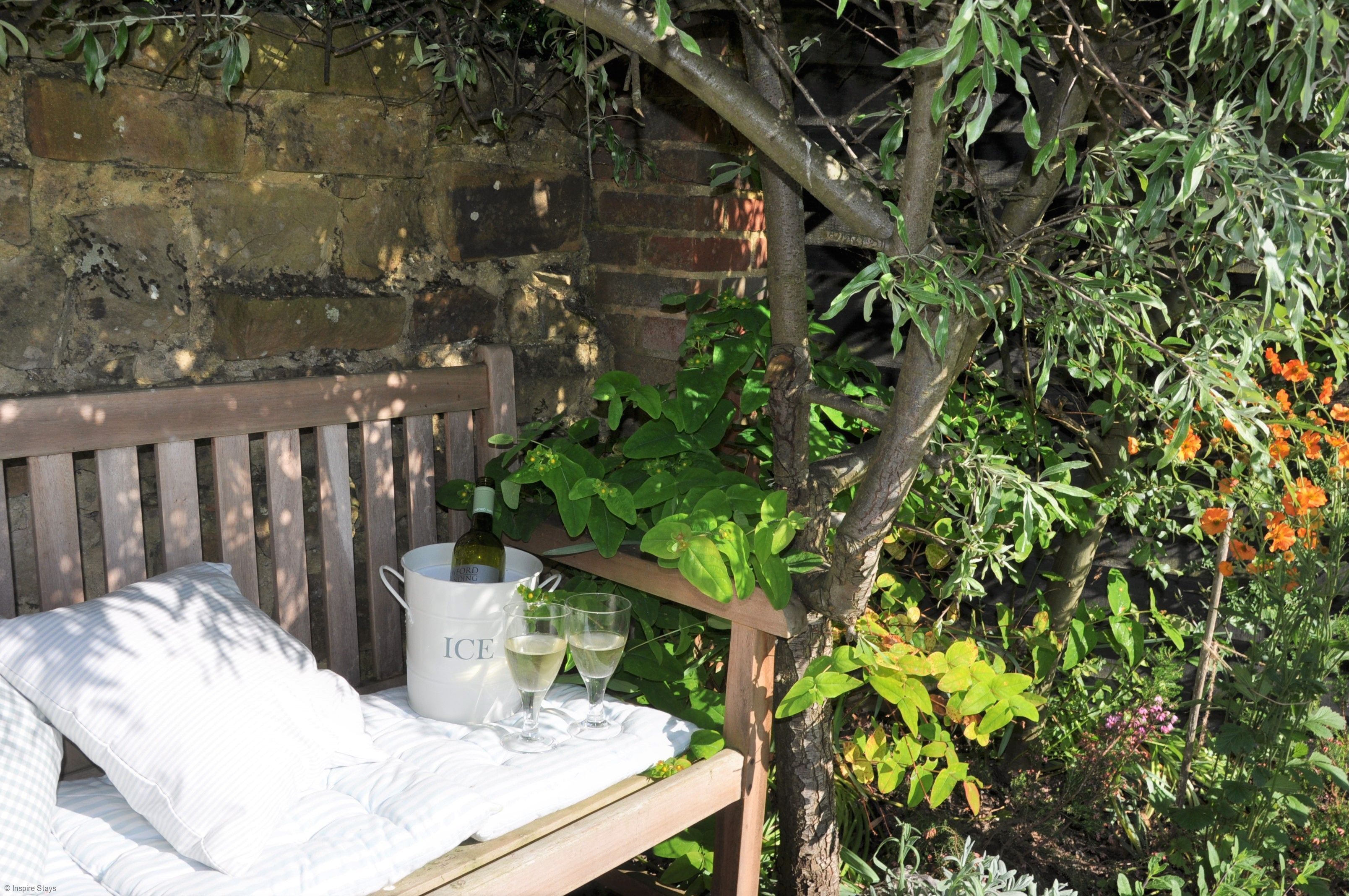 A wooden bench with a white cushion and a white bucket labeled "ICE" containing a bottle and two glasses.
