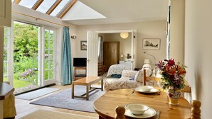 Une pièce spacieuse dotée d’une grande verrière, d’un coin repas avec une table et des chaises en bois, d’un lit confortable agrémenté d’une literie à motifs floraux, et offrant une vue sur un jardin luxuriant grâce à des portes ouvertes.