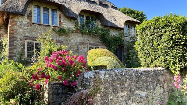 Un bâtiment en pierre avec un toit de chaume, des murs couverts de lierre et un grand rocher au premier plan.