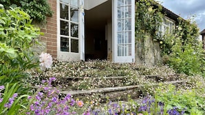 Un jardin où s’épanouit une grande variété de fleurs, avec un escalier en pierre menant à une maison aux portes et fenêtres blanches.