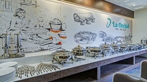 Montaje de bufé con diferentes recipientes de comida y utensilios, un mural con ilustraciones de alimentos y el nombre "La Orchidia" en la pared.