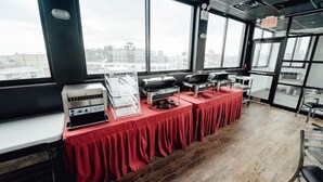 A buffet setup with a red tablecloth, buffet stations, and a view of the city through large windows. - The Feather Factory (Long Island City)