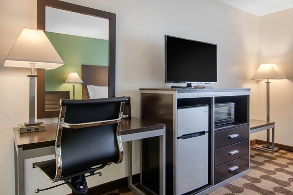 A hotel room with a desk, a chair, a television, a microwave, and a refrigerator.
