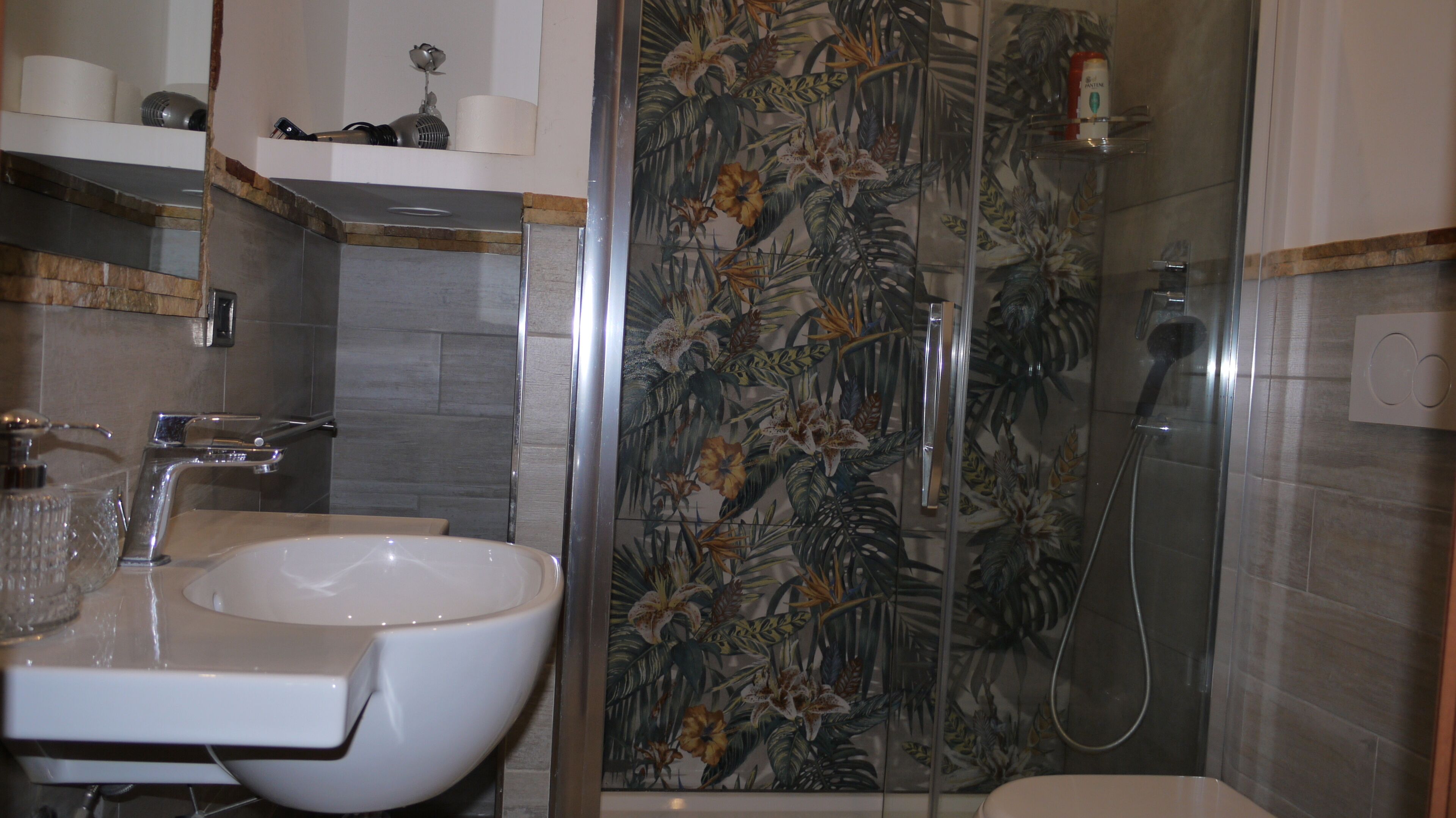 Un baño con un lavabo blanco, una ducha con puerta de cristal y una pared de azulejos con motivos de hojas tropicales.