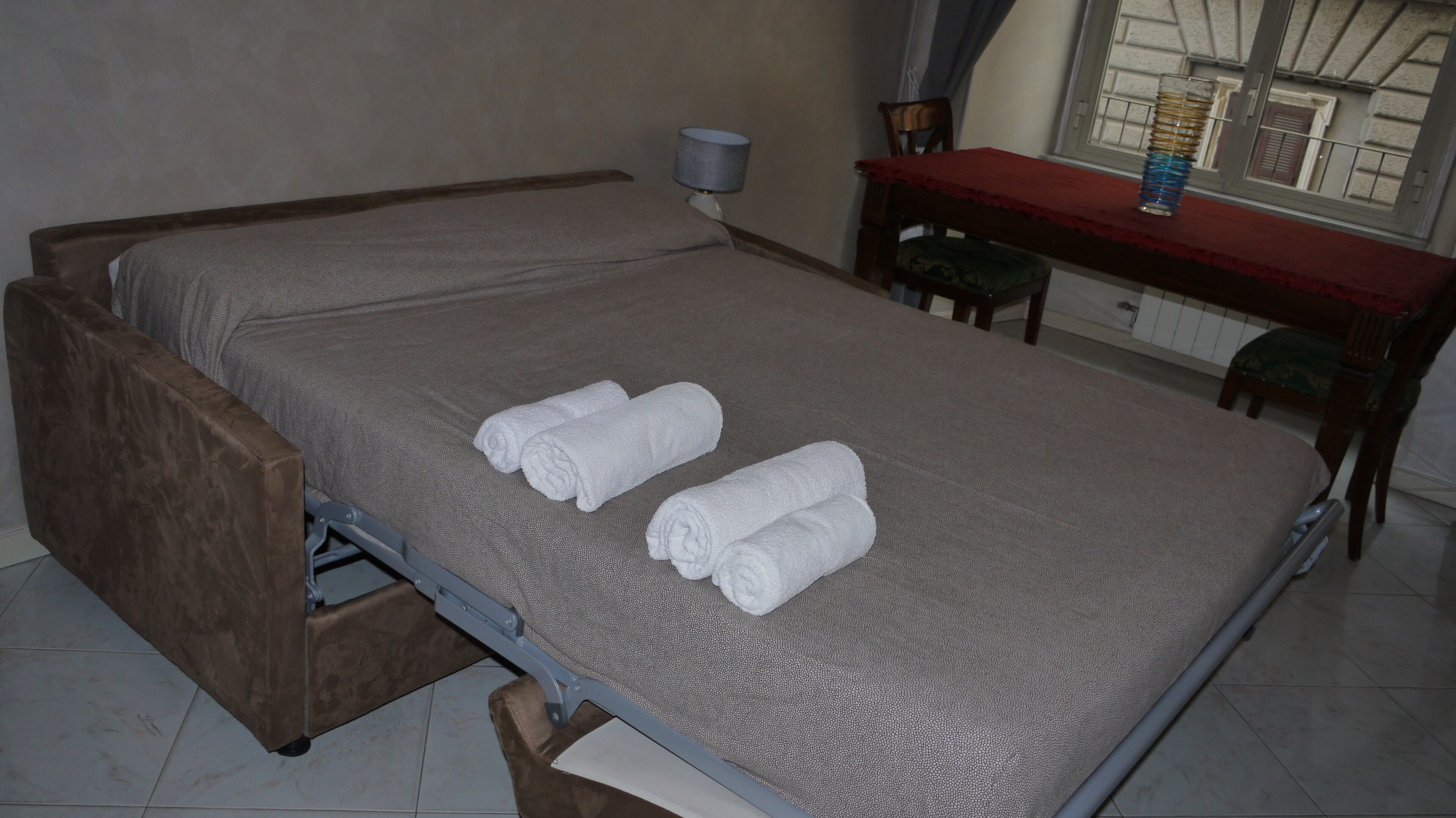 A single bed with a grey mattress and two rolled white towels placed on it.