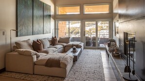 A modern living room with a sectional sofa, a coffee table, and a fireplace. There are large windows providing a view of the outdoors.