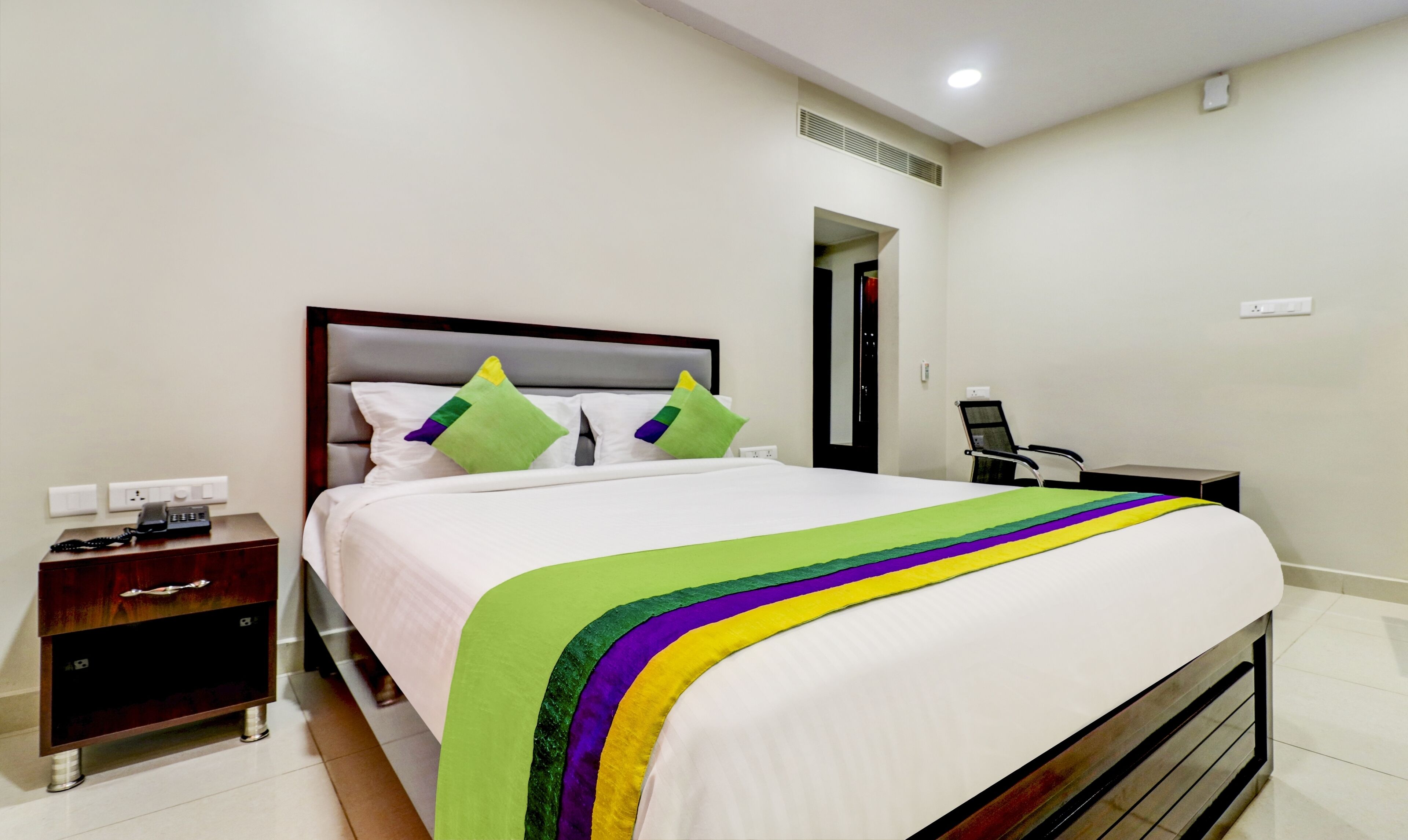 A neatly made bed with a white comforter and colorful pillows, a bedside table with a phone, and a chair in a hotel room.