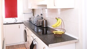 A compact kitchen with white cabinets, a black countertop, a red towel hanging on the wall, a toaster, a kettle, and a banana basket. - Canning Town Cozy one Bedroom Apartment (London)