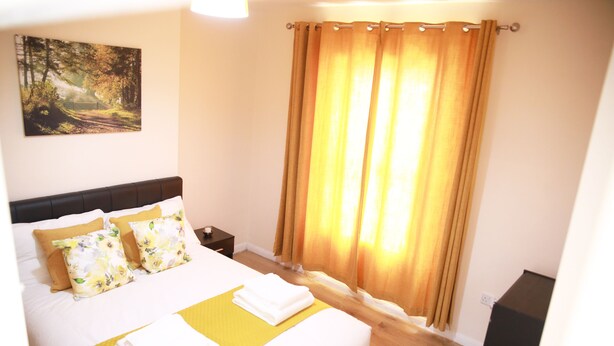 A neatly made bed with white and yellow bedding, a nightstand with a lamp, and a painting of a forest on the wall. - Canning Town Cozy one Bedroom Apartment (London)