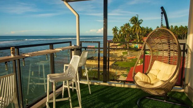 Un balcón con vista al océano, una silla de mimbre y una silla de plástico blanco.
