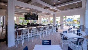 Zona de bar al aire libre con taburetes blancos, un televisor de pantalla plana y vista a la piscina.