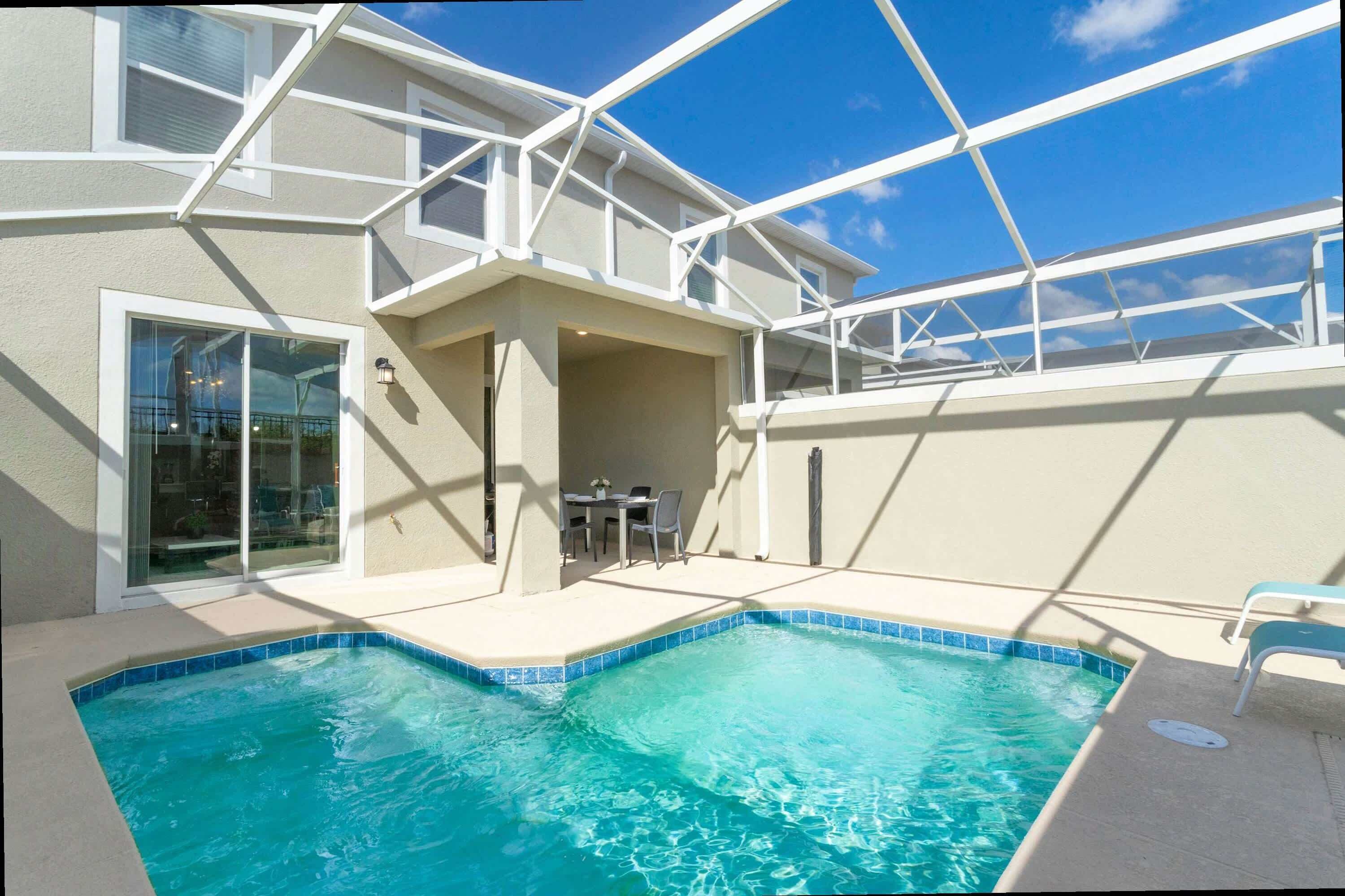 A swimming pool with a clear blue edge, a covered patio area with a table and chairs, and a glass door leading to an outdoor space.
