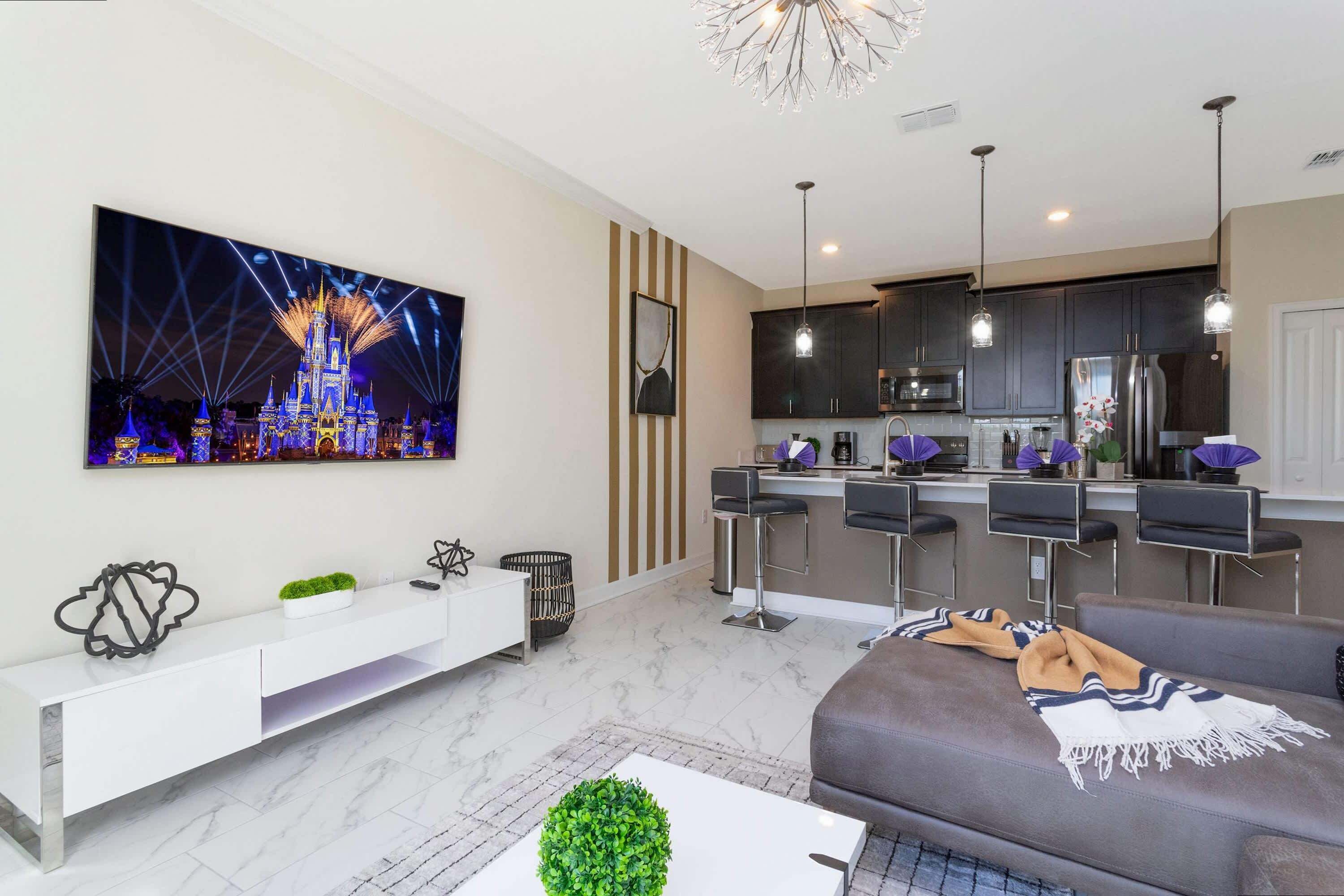 A modern living room with a large flat-screen TV, a sofa, a coffee table, and a kitchen area with a bar.