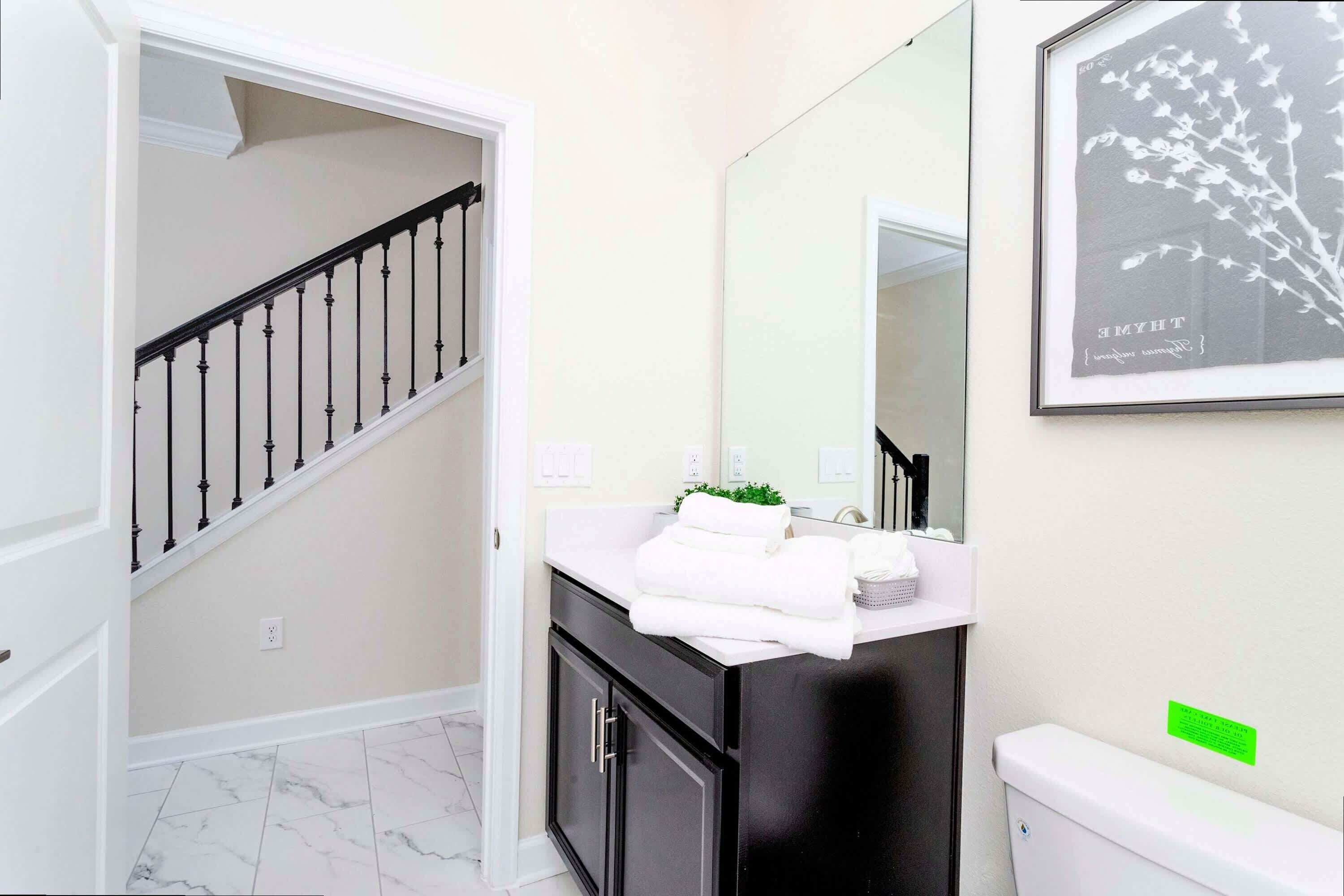 Un baño con inodoro blanco, armarios negros, un lavabo blanco con espejo arriba y una escalera con barandillas negras que conduce a otra habitación.