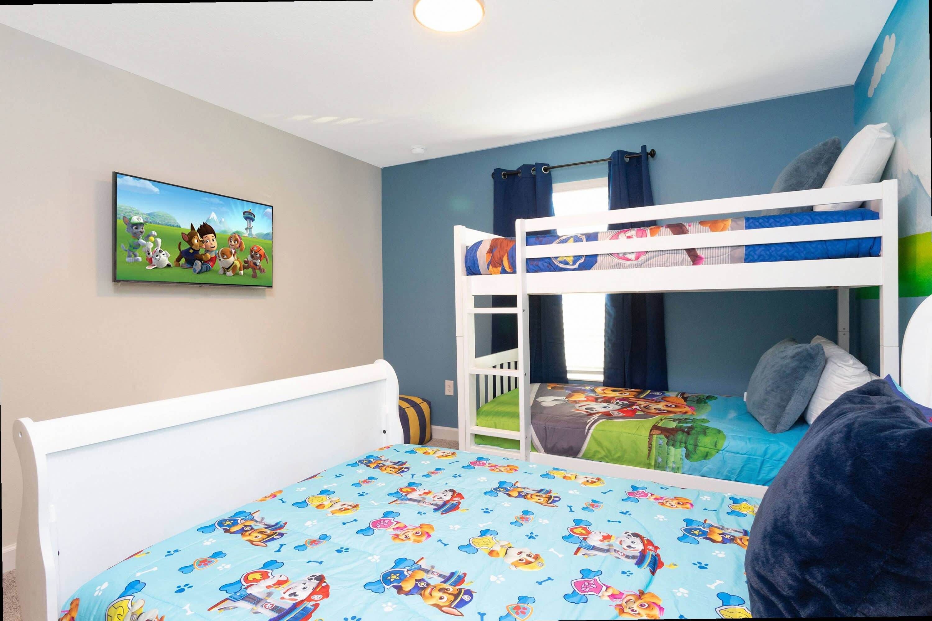 Une chambre d’enfant avec des lits superposés, un téléviseur accroché au mur et un lit recouvert d’une couette aux motifs de dessins animés.