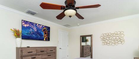 Un dormitorio con una cama grande, ventilador de techo, cómoda, televisor y un adorno en la pared.