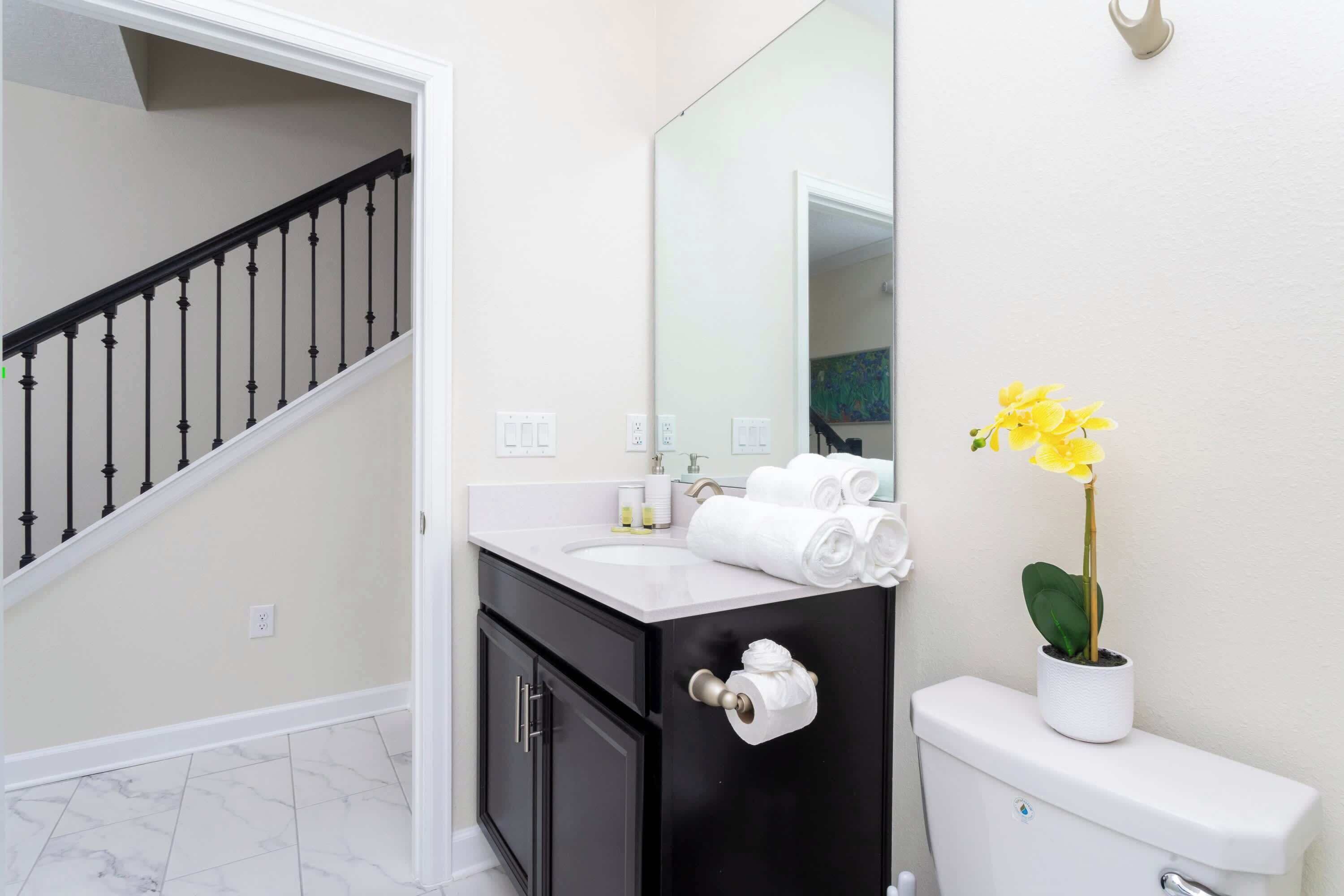 A bathroom with a white sink, black vanity, and a toilet. There is a yellow flower on the toilet and a towel rack with towels.